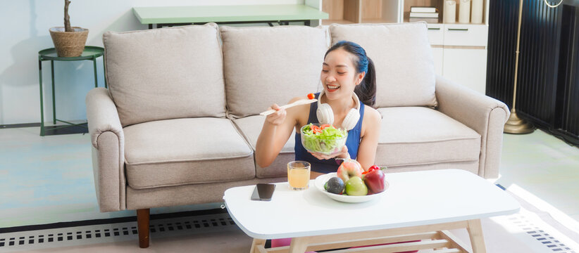 A young Asian housewife enjoys a healthy lifestyle at home, balancing fitness routines with relaxation. She sits on a modern sofa, eating a fresh salad and fruit while embracing healthy habits. - Powered by Adobe