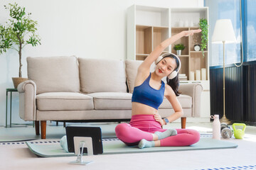 Fototapeta premium A young Asian housewife sits on sofa in her living room, using fitness app on her mobile with headphones. dumbbells and whey protein, she balances yoga, workouts, vegetarian diet and weight loss.