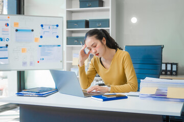 A young Asian businesswoman works alone at a desk with a laptop, stack of papers, and documents....