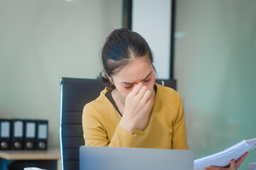 A young Asian businesswoman works alone at a desk with a laptop, stack of papers, and documents. She looks serious and stressed, analyzing financial losses and equity management under daylight.