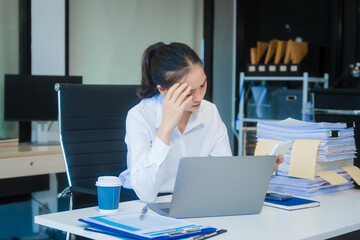 A young Asian businesswoman works alone at a desk under daylight with a laptop, papers, and documents. She appears stressed and serious, dealing with financial reconciliation and variance analysis.
