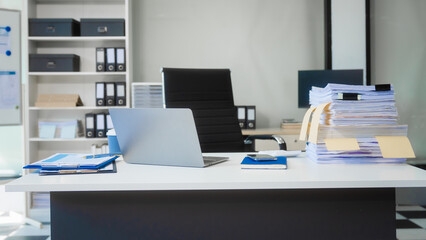 A modern office workspace with a white desk, laptop, stacked papers, clipboard, and document files. Financial charts are spread out under daylight in a clean, bright indoor environment.
