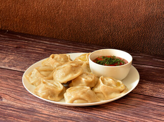 A plate of freshly steamed manti and a cup of tomato spicy sauce on a wooden table.