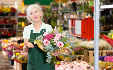 Smiling polite elderly woman in apron working at flower market, offering floral arrangement with fresh gerberas and roses