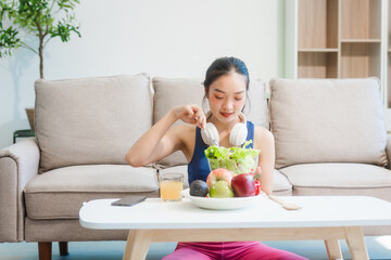 A young Asian housewife enjoys a healthy lifestyle at home, balancing fitness routines with relaxation. She sits on a modern sofa, eating a fresh salad and fruit while embracing healthy habits.