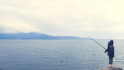 A lone child stands by the lake, fishing under a misty sky.