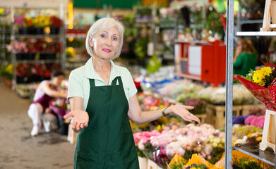 Glad aged woman seller posing against the background of colorful plants in point of sale of flowers