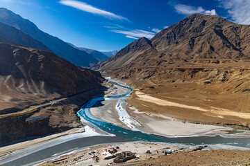 Panoramic view of confluence of river Indus and river Zanskar at Sangam point with mountains, sky and clouds in the background.