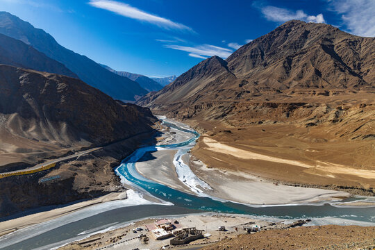 Panoramic view of confluence of river Indus and river Zanskar at Sangam point with mountains, sky and clouds in the background.