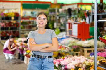 Young woman in casual wear, confident owner of floral shop standing with crossed arms among stall with colorful blooming cut and potted flowers, looking at camera with smile
