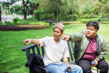 Obraz premium Two College Man Sitting on Bench and Talking While Showing Something By Pointing Index Finger. 
