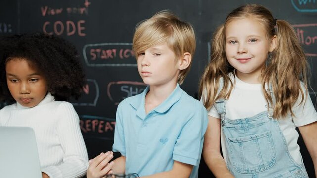 Smart girl using laptop programing engineering code with diverse friends ay table with microchip and main board scatter around in STEM classroom at blackboard written with coding prompt. Erudition.