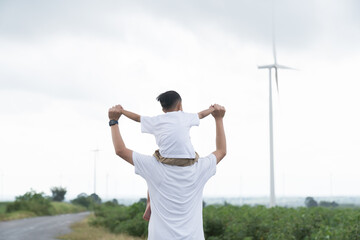 Happy Asian family, father carrying son on shoulder while playing together at wind turbine farm...