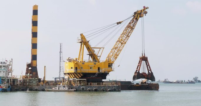 large, yellow floating crane on a barge in a body of water. The crane has a long boom with multiple cables and is equipped with a clamshell bucket, used for dredging.