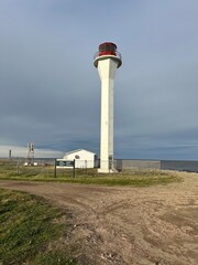 A tall, white lighthouse with a red lantern room stands near the coast, surrounded by a fence. A small building and a sign are visible at the base, with a dirt path leading up to it.
