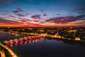 Obraz premium Vibrant sunset colors reflect on serene water as twilight descends over a city adorned with lights