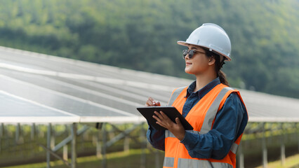 Female electrical engineer wearing hard hat inspecting solar farm panel equipment. Eco-friendly...