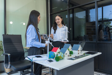 Two businesswomen sit at a table in the office, discussing a model village with windmills,green...