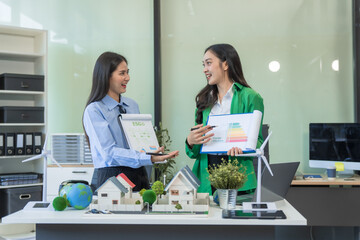 Two businesswomen sit at a table in the office, discussing a model village with windmills,green...