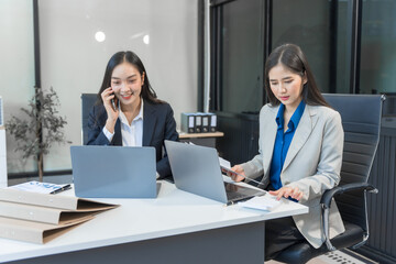 Two Asian businesswomen work on a laptop in an office, pointing at the screen, talking, and smiling with joy as they celebrate positive results of their business success.