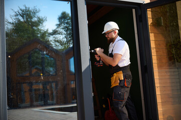 Construction worker installing door in a new house construction site. Man in working attire install front door. Builder in uniform with screwdriver adjusting front glass door in new beautiful house.