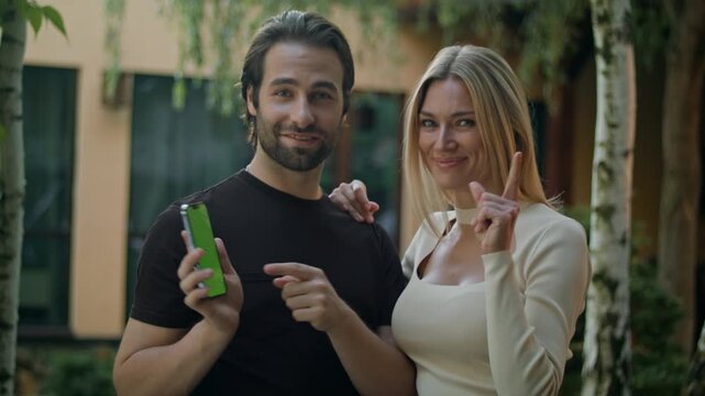 Outside, the couple taps their smartphone in a mobile app, winning a prize. Man turns the green screen camera-facing, pointing at it to highlight the app&rsquo;s excellence, celebrating their success.