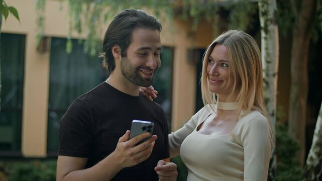 Standing outside, they tap the smartphone screen and win a prize on a mobile app. Man shows the green screen to the camera, proudly pointing at it, celebrating their big victory and success.