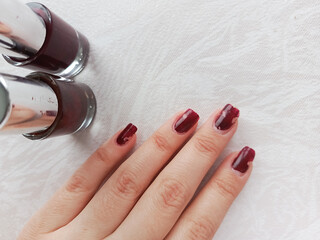 Chipped burgundy nail polish. A woman's hand with a ruined manicure and a bottle of red nail polish on the table.