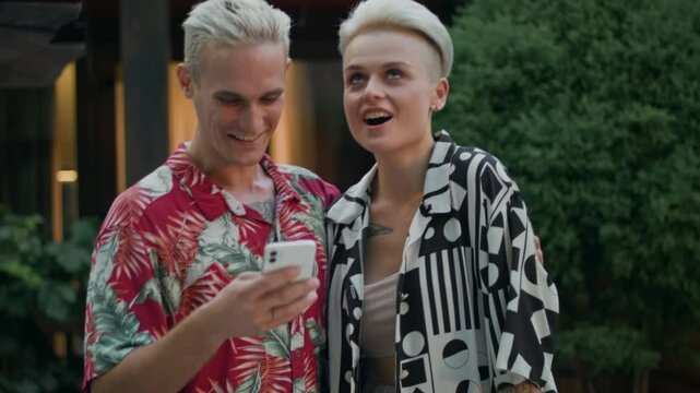 A blonde guy and girl with tattoos hold a phone outside. They look at the screen and win a prize in a mobile app. Overjoyed, they can't contain their emotions. He hugs his girlfriend.