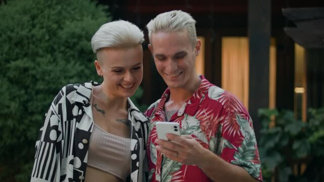 A blonde man and woman with tattoos hold a phone outdoors. They check the screen and win a prize in a mobile app. Elated, they cannot contain their emotions. He joyfully hugs his girlfriend.