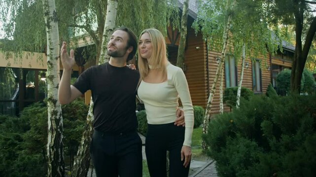 A man hugs his girlfriend by the waist as they stroll past a country house. Amid beautiful landscaping and nature, he points to something ahead, and they head in that direction.
