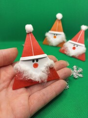 Handmade paper Santa Claus decorations with cotton beards are shown against a green background. One is held in a hand, with others placed in the background, creating a festive and crafty vibe.