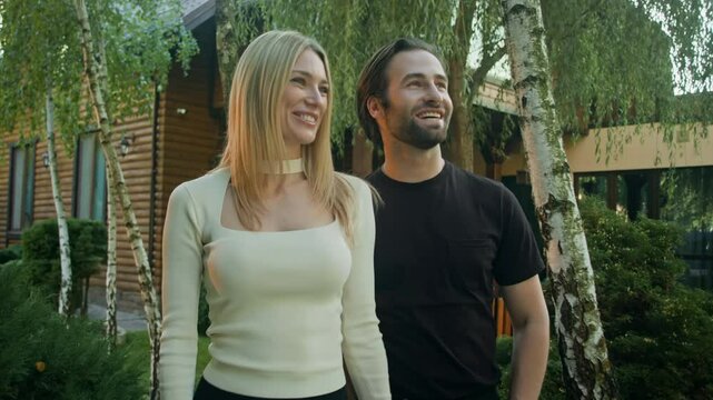 A blonde woman and a brunette man, a loving couple, walk around a country vacation resort. He gently wraps his arm around her waist and softly kisses her on the lips.