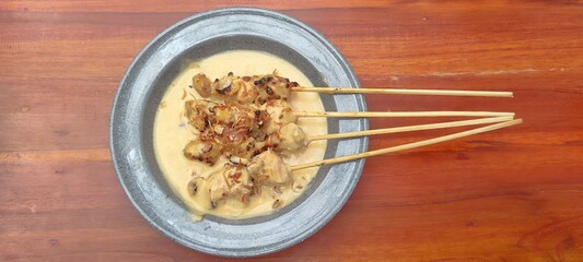 a plate of grilled chicken satay serepeh, served with additional coconut milk sauce, sliced ​​shallots, chopped red chilies, and halved lime, on a gray plate, isolated grey plate, wooden table