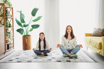 Relaxed young mother and daughter meditating together in cozy living room embracing natural light and tranquility on a beautiful spring day