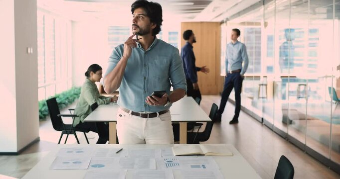Middle-aged Indian manager, confident business leader using smartphone to message. Busy professional works on business process map in corporate office. Communication and strategy planning for success