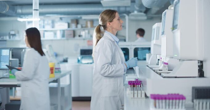 Team of Biomedical Engineers Work in a Lab. Senior Researcher Adding Samples to Test Machine, Asian Female Adjusting Settings on a Digital Microscope while Male Colleague Carrying Medical Glassware