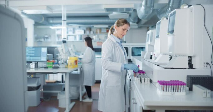 Biomedical Researchers Conduct Experiments in a Modern Laboratory. Female Scientist Preparing a Set of Samples in Test Tubes with Purple Caps. Colleague Analyzes Data on a Computer in the Background