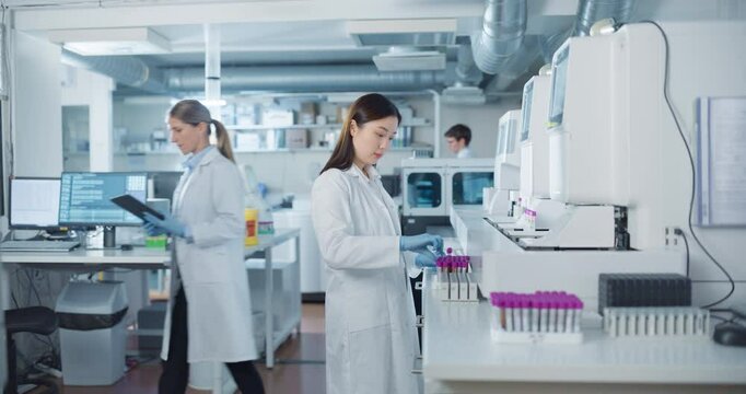 Team of Young Biochemists Collaborating in a Laboratory. Asian Female Researcher Loading Test Tubes with Blood Samples Into a Centrifuge while Her Coworker Reviews Experimental Data on a Computer
