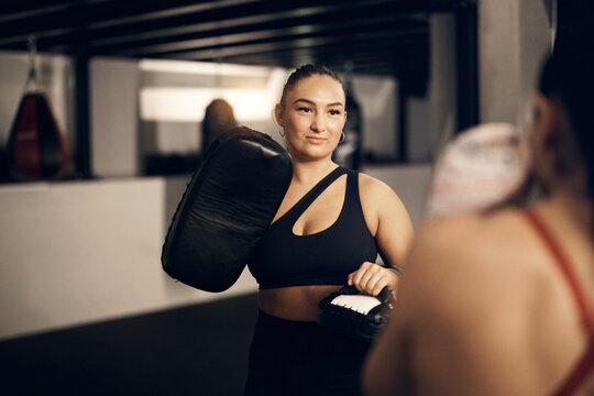 Fit young woman in sportswear smiling while holding pads for a female partner practicing punching during a boxing workout in a gym