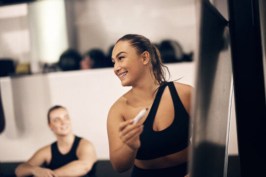 Smiling female coach drawing exercise examples on a whiteboard for a group of fit young woman during a break for a workout class in a gym