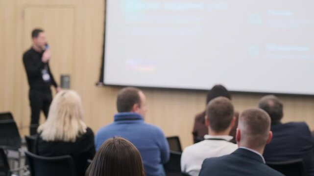 Conference speaker presenting to audience in seminar. Attendees focused on technology presentation, engaging atmosphere, professional setting.