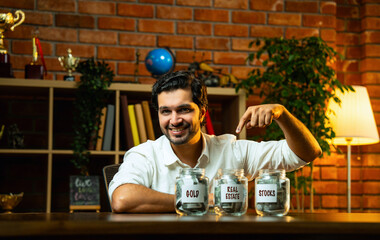 Indian bearded young man sitting at home at desk saving money in glass jars