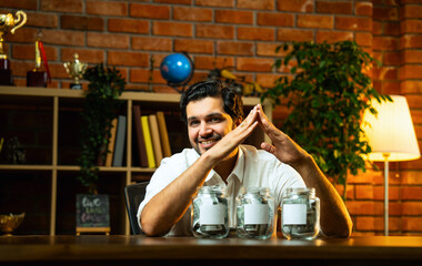 Indian bearded young man sitting at home at desk saving money in glass jars