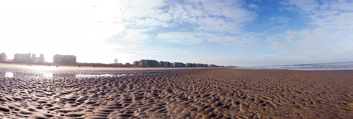 A panoramic view of a vast sandy beach with a city skyline under a bright blue sky. The tranquil scene evokes peacefulness and escape, perfect for a background or travel concept.