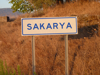 Blue 'Sakarya' Sign on a Metal Pole: A Bold Contrast Against the Natural Landscape of Brown Grass and Soft Shadows, Capturing a Serene Afternoon in the Heart of the City.