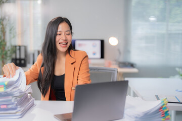 Fototapeta premium Asian businesswoman is smiling and holding documents while having a video conference on her laptop in a bright office, she is wearing an orange blazer and a black shirt