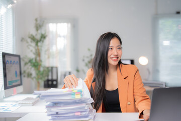 Smiling asian businesswoman managing paperwork and using laptop, sitting at desk in modern office, working with financial charts and graphs, enjoying her productive job