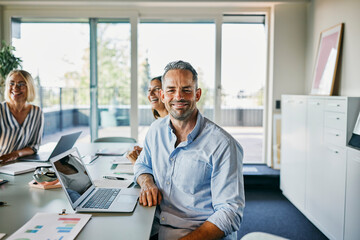 Happy Business Team Meeting in a Modern Office Environment. A mature businessman, with a hint of gray in his hair, wearing a blue shirt, looks directly into the camera.
