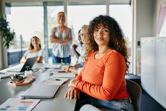 Confident Businesswoman Leading a Creative Team in a Modern Office Setting. Her colleagues stand behind her, looking into the camera, smiling.